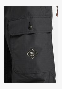 Pantalones cargo negros con un patrón de cuadrícula texturizado. Presenta un gran bolsillo frontal con cierre de botón y un parche de logo de la marca DC.