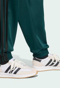 Pantalons de sport verts avec des rayures latérales noires, associés à des baskets blanches avec des rayures noires et une semelle en caoutchouc. Manches élastiques visibles.