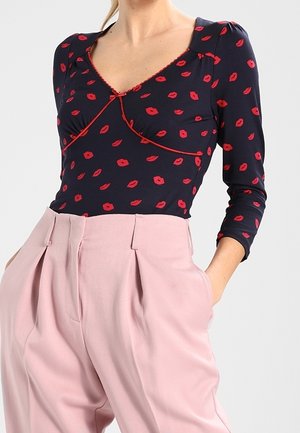 Vrouw draagt een marineblauwe top met lange mouwen met rode lippenafdrukken en hooggetailleerde, geplooide lichtroze broek met handen in de zakken.