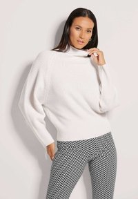 Weißer Strickpullover mit hohem Kragen und lockerer Passform, ausgestattet mit strukturierten Details. Kombiniert mit schwarzen und weißen gemusterten Leggings.