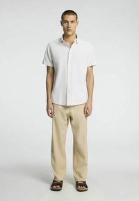 Kortärmad vit skjorta med knappar och ett strukturerat mönster, matchad med beigefärgade löst sittande byxor och svarta sandaler. Modellen står framåtvänd.