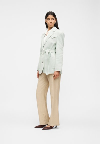 Γυναίκα με ανοιχτό πράσινο σακάκι με ζώνη, λευκό πουκάμισο, μπεζ παντελόνι με φαρδιά μπάκα και κουμπιά κοντά στους αστραγάλους και καφέ μυτερά τακούνια, στέκεται μπροστά σε λευκό φόντο.