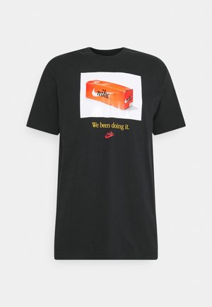 Czarna koszulka z pomarańczową grafiką pudełka na buty Nike i żółtym tekstem z napisem "We been doing it" nad czerwonym logo Nike.