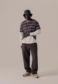 Μπλούζα κοντομάνικη με μοβ και λευκές ρίγες, φορεμένη πάνω από μια λευκή μακρυμάνικη μπλούζα, συνδυασμένη με σκούρο παντελόνι και μπεζ αθλητικά παπούτσια. Περιλαμβάνεται καπέλο τύπου bucket.