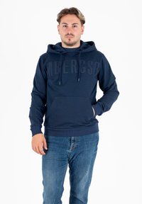 Felpa con cappuccio blu navy con logo testurizzato sulla parte frontale, cappuccio con coulisse e tasca a marsupio. Abbinata a jeans blu chiaro, il tessuto appare morbido.