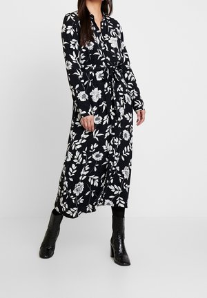 Vrouw draagt een zwarte midi-jurk met lange mouwen en witte bloemenprint, met knopen aan de voorkant, een riem in de taille, zwarte legging en zwarte laarzen met hak.