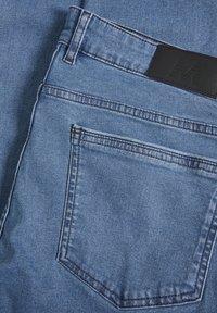 Vaqueros de mezclilla azul con un bolsillo con parche y una etiqueta de cinturón de cuero negro. La tela tiene una textura suave y un detalle de costura clásico.