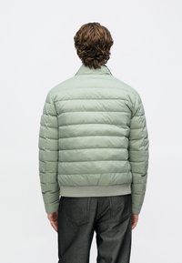 Casaco de penas verde-claro com design estruturado, costuras horizontais e punhos e bainha canelados, combinado com jeans escuros de ganga.