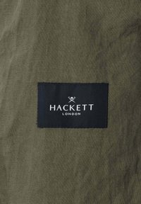 Dunkelgrüner Stoff mit einem aufgenähten schwarzen rechteckigen Etikett, auf dem „HACKETT LONDON“ in weißer Schrift und ein kleines, gekreuztes Polomallette-Logo zu sehen sind.