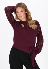 Burgundin värinen kietaisuneule, jossa on ribbikuvio, pitsiset pitkät hihat; sisältää vyötärönsidontayksityiskohdan ja avorintaleikkauksen. Yhdistetty mustiin housuihin.