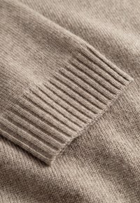 Beige strikket stof med ribbet tekstur langs kanterne, der har et blødt og fint udseende, som viser en glat og jævn vævning.