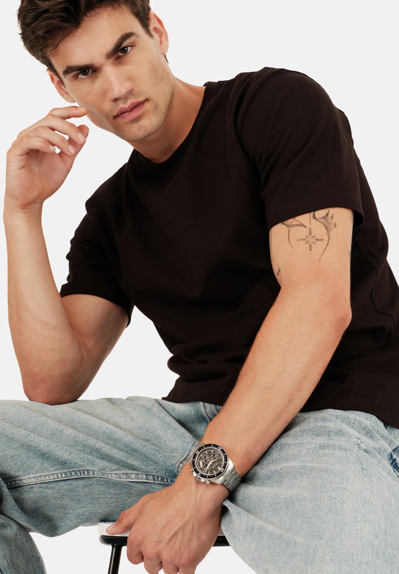 T-shirt noir à manches courtes, associé à un jean en denim bleu clair, complète une montre-bracelet argentée avec un cadran noir et des détails de chronographe.