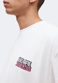 Κοντινό πλάνο ενός ατόμου που φοράει λευκό πουκάμισο με το κείμενο «WE ARE JUVENTUS» τυπωμένο στο στήθος με μαύρα και ροζ γράμματα.