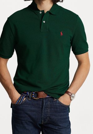 Mężczyzna w ciemnozielonej koszulce polo z krótkim rękawem z czerwonym logo, ciemnych dżinsach, pasku w paski i zegarku na rękę z brązowym skórzanym paskiem. Ręce w kieszeniach.