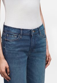 Femme portant un jean en denim bleu et un haut sans manches blanc en côte, les mains reposant le long du corps.