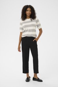 Woman with curly hair wearing a gray and white striped short-sleeve shirt, black cropped pants, and black loafers, standing with one hand in pocket.