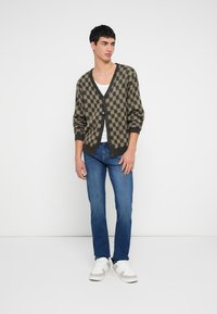 Cardigan s tmavě zeleným a béžovým kostkovaným vzorem, s dlouhými rukávy a černými knoflíky, ve spojení s modrými džínami a bílými teniskami.