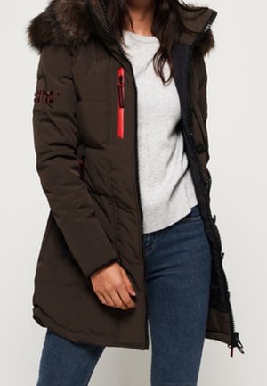 Woman wearing dark brown long winter coat with fur hood, red zipper accents, gray sweater, and blue jeans.