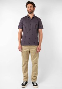 Polo à manches courtes violet foncé avec un imprimé de palmiers noirs, associé à un chino beige et des baskets noires avec des semelles blanches.