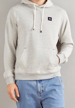 Person iført en lysegrå hættetrøje med frontlomme og hvide snore, kombineret med lyseblå denimjeans, stående mod en ensfarvet baggrund.