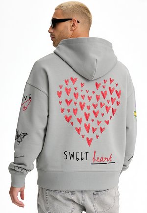 Man with short blonde hair and sunglasses wearing a gray hoodie with red hearts forming a large heart and "SWEET heart" text on the back.