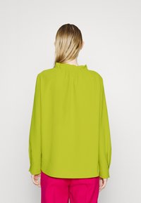 Limegrøn bluse med rynket krave, lange ærmer og løs pasform, parret med lyserøde bukser. Glat tekstur, minimal detaljer.