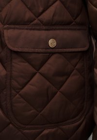 Κοντινό πλάνο τσέπης καφέ μπουφάν με καπιτονέ σχέδιο, με καπάκι ασφαλισμένο με στρογγυλό χρυσό κουμπί που φέρει λουλουδένιο έμβλημα.