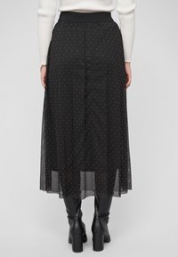 Sort, gennemsigtig lang nederdel med prikket mønster, elastisk taljebånd og lagdelt design. Båret med knælange sorte støvler.