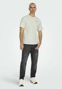 White cotton t-shirt with small red logo, paired with dark grey jeans. Footwear is beige sneakers with green accents. Simple, casual outfit.