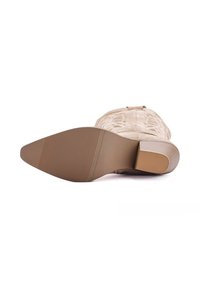 Bota de vaquero de ante beige tumbada de lado, mostrando la suela texturizada y el tacón cuadrado con apariencia de madera apilada.