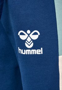 Blauwe sportkleding van een gestructureerde stof, met een wit logo en de merknaam "hummel" prominent op de zijkant gedrukt.