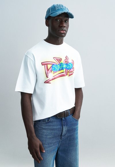 Λευκό t-shirt με κοντά μανίκια και πολύχρωμο τύπωμα σε στυλ γκράφιτι, συνδυασμένο με μπλε τζιν παντελόνι και ανοιχτό μπλε τζιν καπέλο.