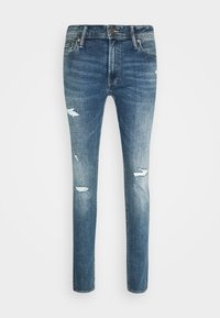 Jeans de corte slim en un denim azul desgastado con detalles desgastados y ligeras deshilachaduras en las rodillas. Cuentan con un cierre frontal de botón y cremallera.