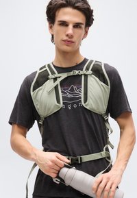 Νέος άνδρας που φοράει πράσινο γιλέκο πεζοπορίας πάνω από μαύρο t-shirt με γραφικά, κρατώντας με τα δύο χέρια ένα γκρι θερμομονωτικό μπουκάλι νερού.