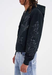 Felpa con zip nera con grafiche grigie, caratterizzata da motivi di aquila e teschio, insieme a elementi testuali. Materiale in cotone morbido.