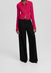 Camisa ajustada de canalé rosa con botones en la parte delantera, combinada con pantalones vaqueros negros de pierna ancha. Textura de tejido suave y detalles de diseño elegantes.