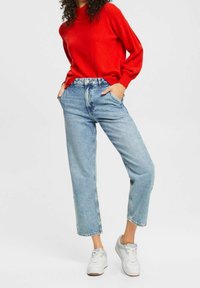 Frau trägt einen hellroten Pullover mit langen Ärmeln, hellblaue High-Waist-Jeans und weiße Turnschuhe, steht mit den Händen in den Taschen.