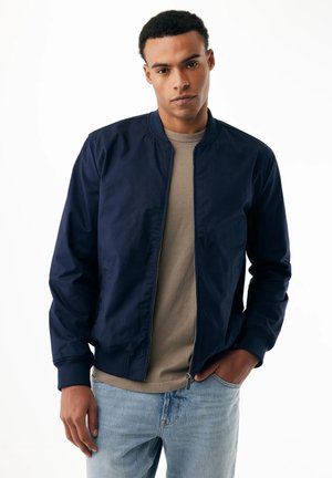 Homme portant une veste bomber bleu marine sur un t-shirt beige et un jean bleu clair, debout avec une main dans la poche, sur un fond blanc.