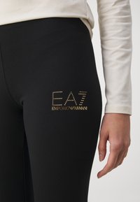 Zwarte leggings van rekbare stof met een gouden studs logo "EA7 EMPORIO ARMANI" op de linker bovenbeen. Gladde textuur, aangesloten ontwerp.