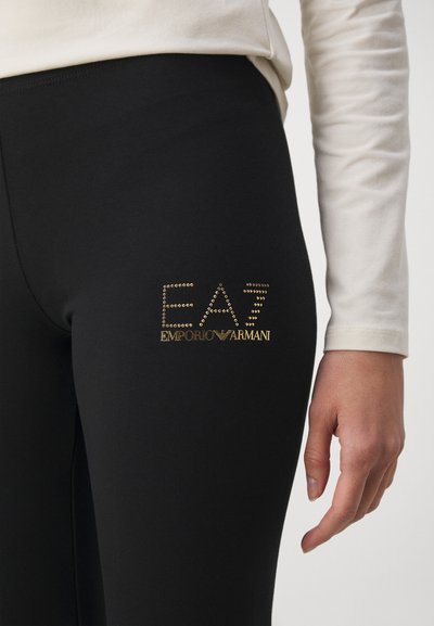 Черни легинси от еластична материя с логото "EA7 EMPORIO ARMANI", украсено с златни пирончета, от лявата страна на бедрото. Гладка текстура, прилягащ дизайн.