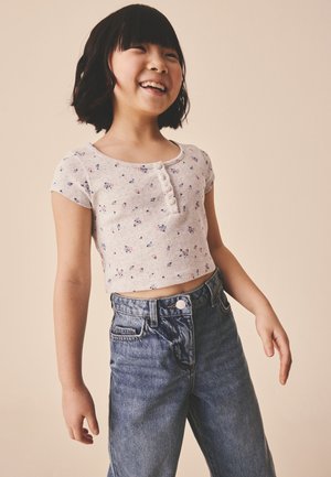 Smilende pige med kort sort hår iført en lys blomsterprintet cropped top og højtaljede blå jeans foran en ensfarvet beige baggrund.