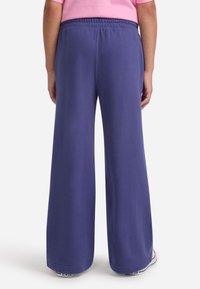 Fialové široké teplákové nohavice s elastickým pásom, mäkkou textúrou a nenápadnými prešívacími detailmi, skombinované s bielymi teniskami.