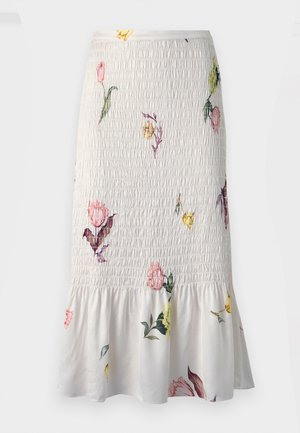 Kvinde iført en kort mørk denimjakke, hvid blomsterprintet midi nederdel med flæsekant og hvide spidse hæle, stående foran en ensfarvet baggrund.