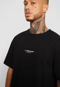 T-shirt de algodão preta com decote redondo, mangas curtas e um grafismo frontal com "DENIM PROJECT" e coordenadas em texto branco e vermelho.