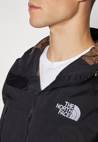 Chaqueta negra con cierre frontal, capucha y forro interior marrón. Cuenta con un logo blanco en el pecho y acentos de tela texturizada.