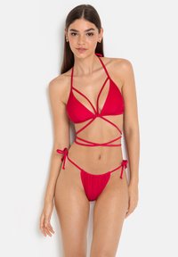 Punainen bikinit, joissa on nauhalla kiinnitettävä halterneck-toppi, jossa on leikkuureikiä, sekä vastaavat matalat alushousut, joissa on solmittavat saumat. Sulava ja joustava materiaali.