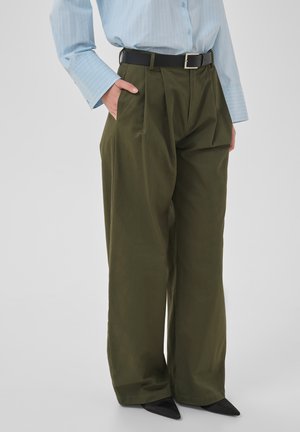 Personne portant un pantalon large olive à plis devant, une ceinture noire, une chemise bleu clair à fines rayures et des chaussures noires pointues.