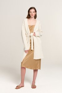 Weißer gestrickter Cardigan mit Bindedetail in der Taille, getragen über einem glänzenden goldenen Nachtkleid. Das Model trägt braune flache Sandalen. Neutraler Hintergrund.