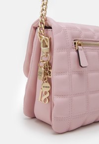 Jasnoróżowa pikowana torba na ramię z złotym okuciem, z dekoracyjnym zawieszką, zamykaną kieszonką oraz łańcuchowym paskiem do noszenia.