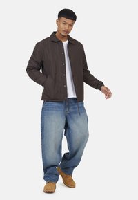 Veste légère marron avec un col, fermeture à boutons-pression et poignets élastiques, assortie à un jean en denim bleu à coupe ample et des chaussures marron.